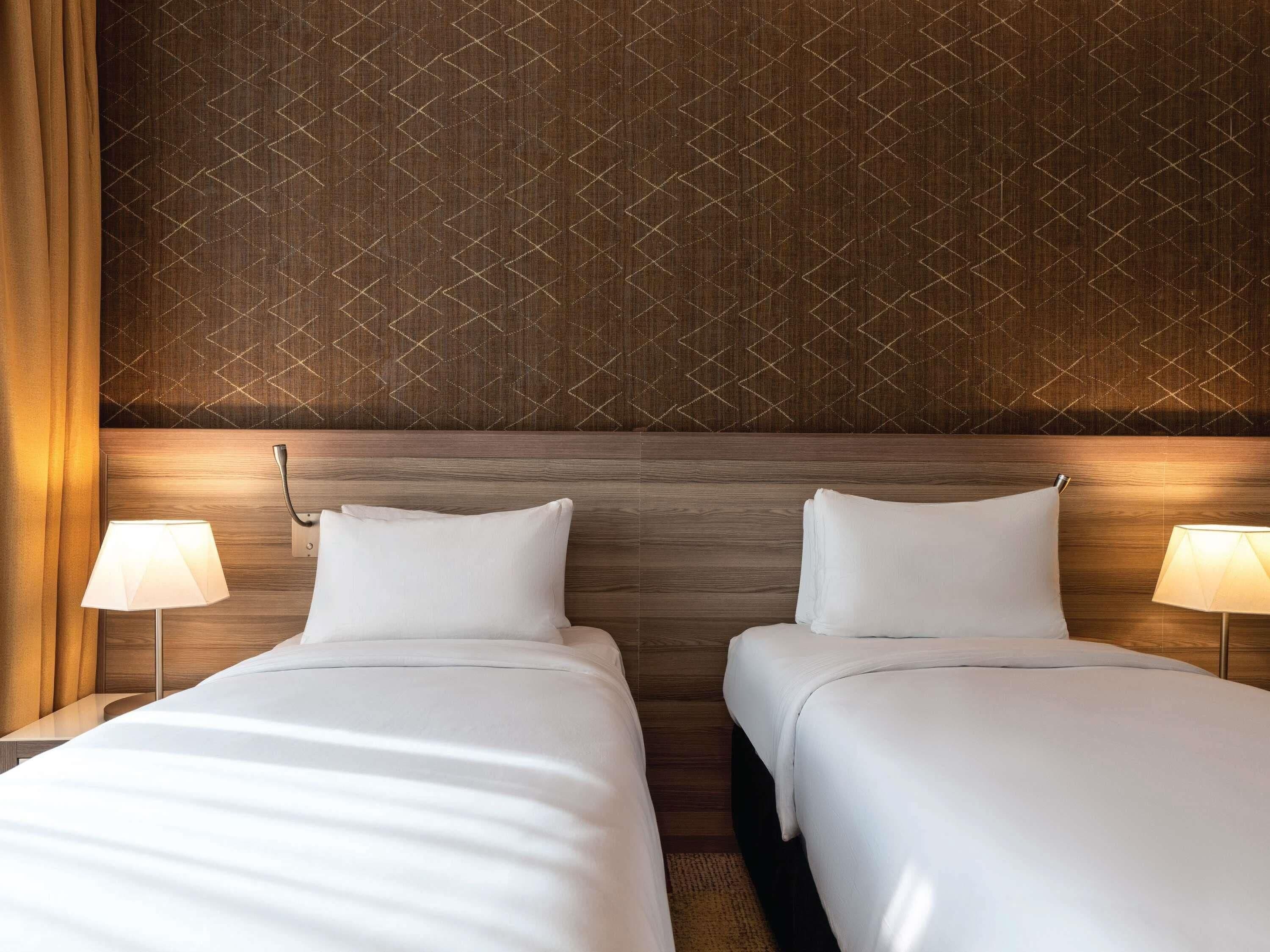 Quarto de hotel com duas camas, cabeceira de madeira, papel de parede estampado e luminárias de cabeceira.