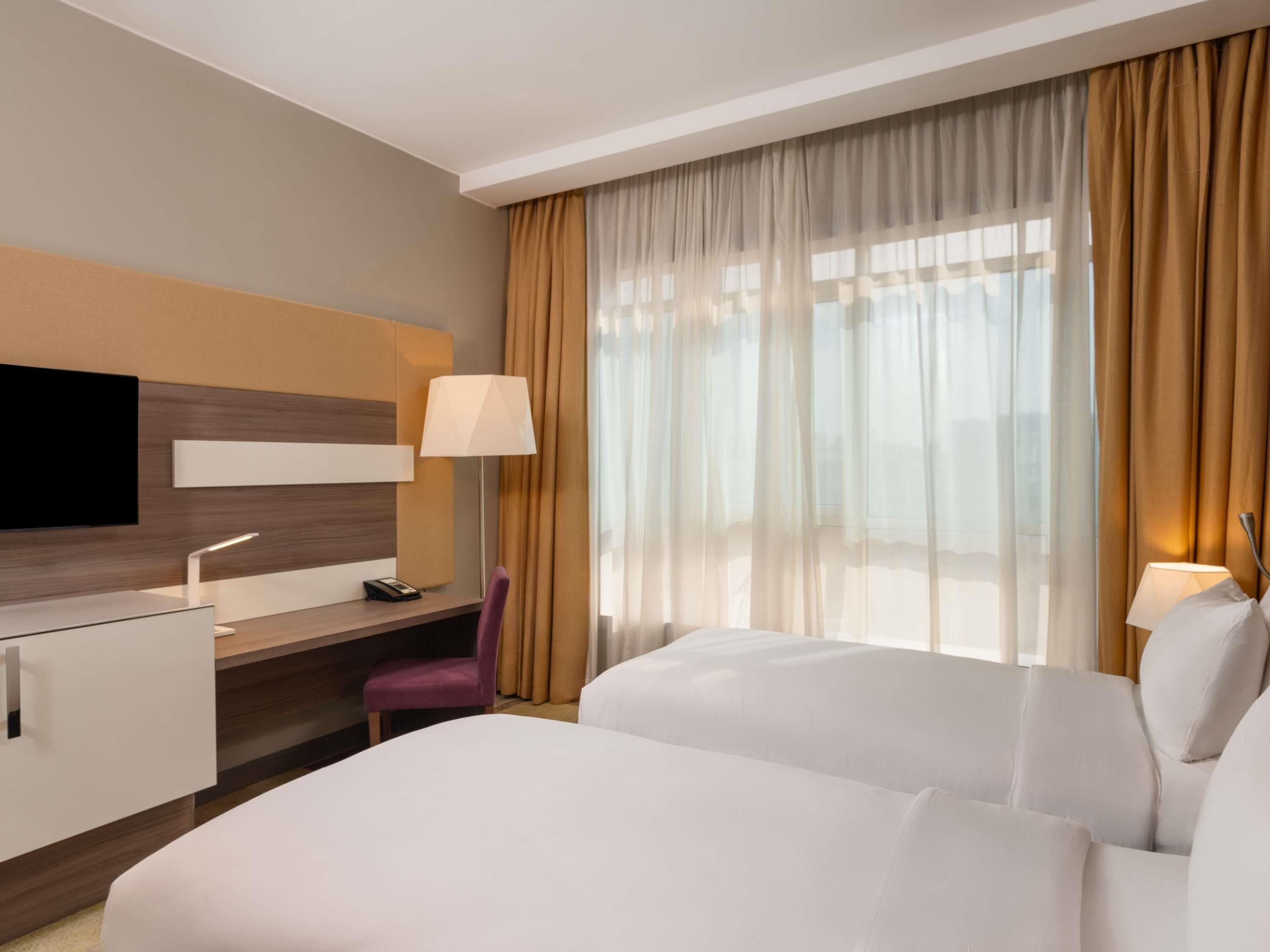 A hotel room with a bed, a desk, a chair, a TV, and a window with curtains.