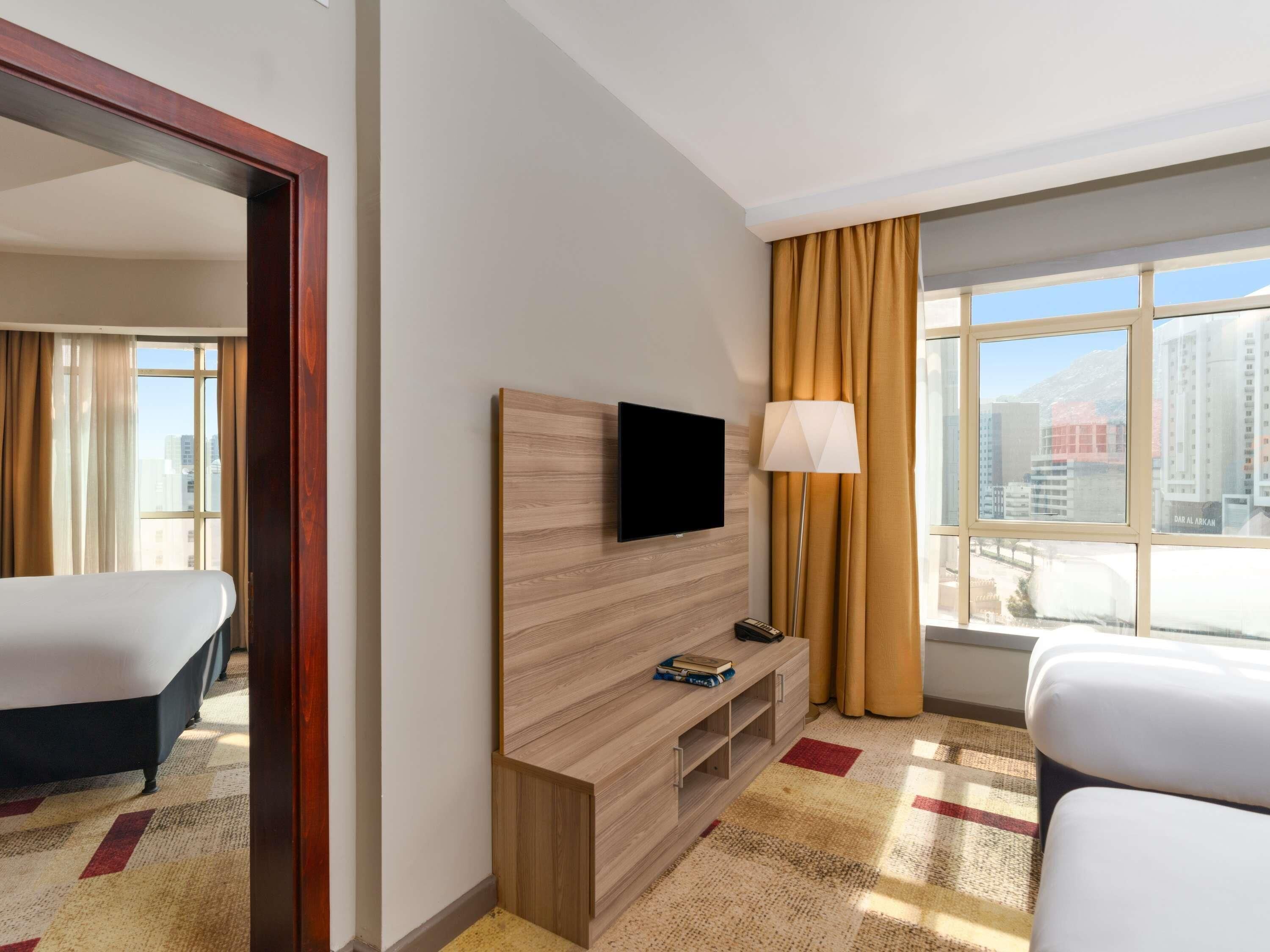 A hotel room with a bed, a TV mounted on a wooden panel, a lamp, and a view of the city through large windows.
