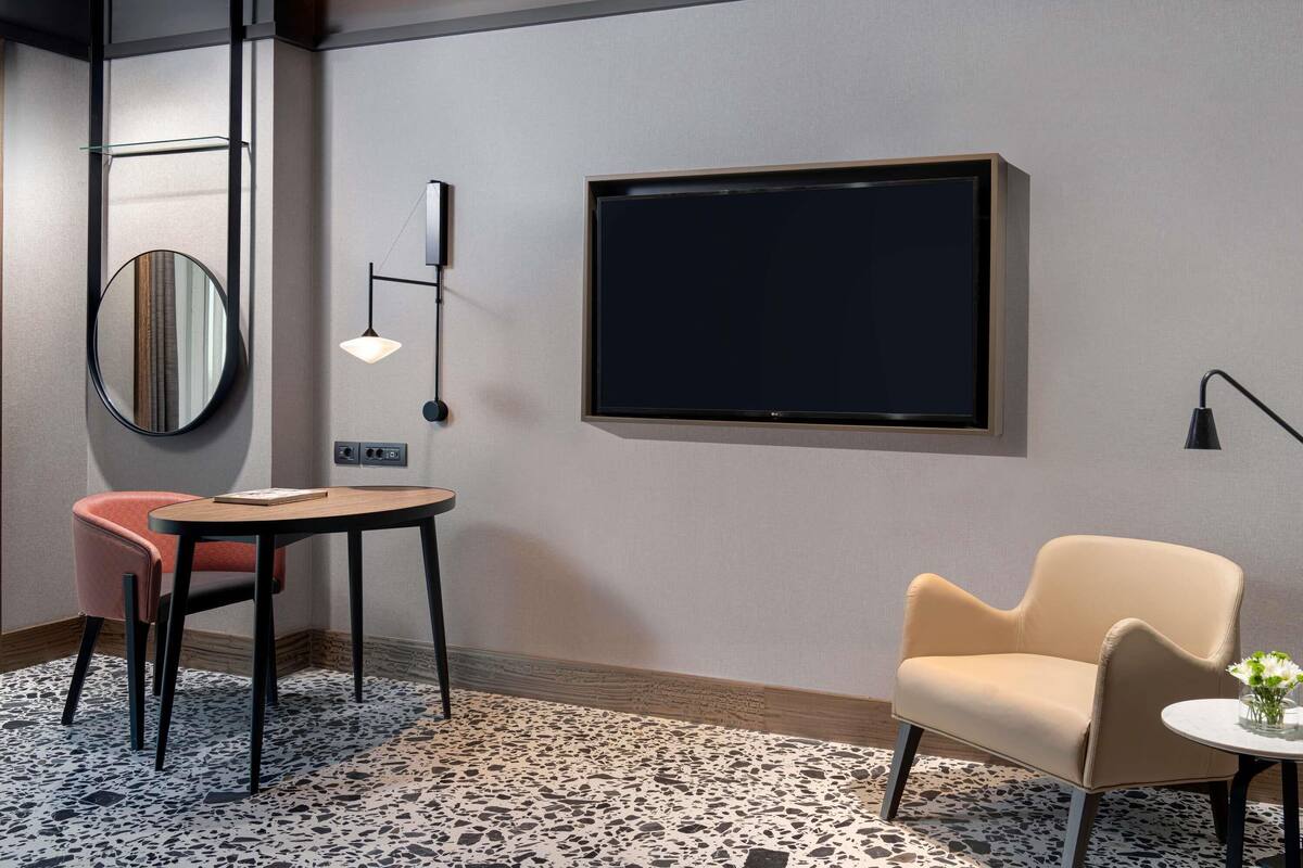 A modern hotel room with a round mirror, a flat-screen TV, a small table, a chair, and a sofa.