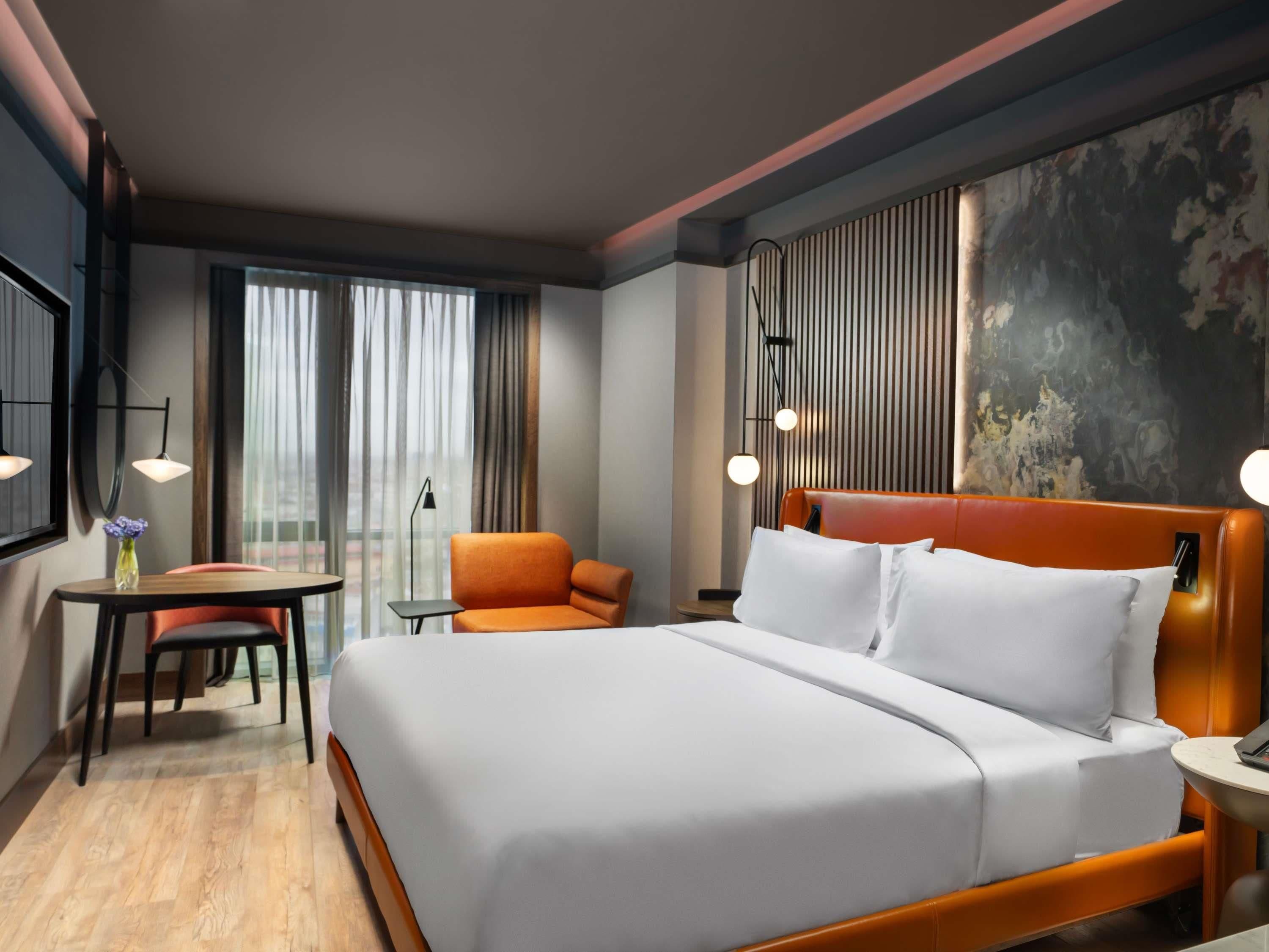 A modern hotel room with a large bed, a small table with a chair, a sofa, and a view of the city through the window.