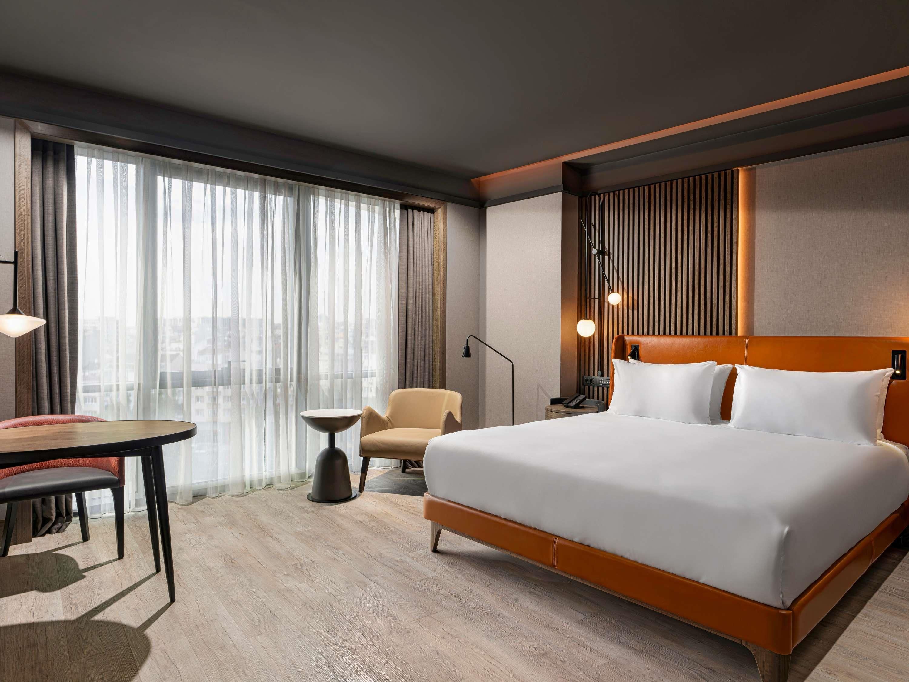 Quarto de hotel com uma cama grande, uma escrivaninha, uma cadeira e vista para a cidade através de cortinas leves.