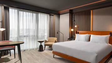 Quarto de hotel com uma cama grande, uma escrivaninha, uma cadeira e vista para a cidade através de cortinas leves.