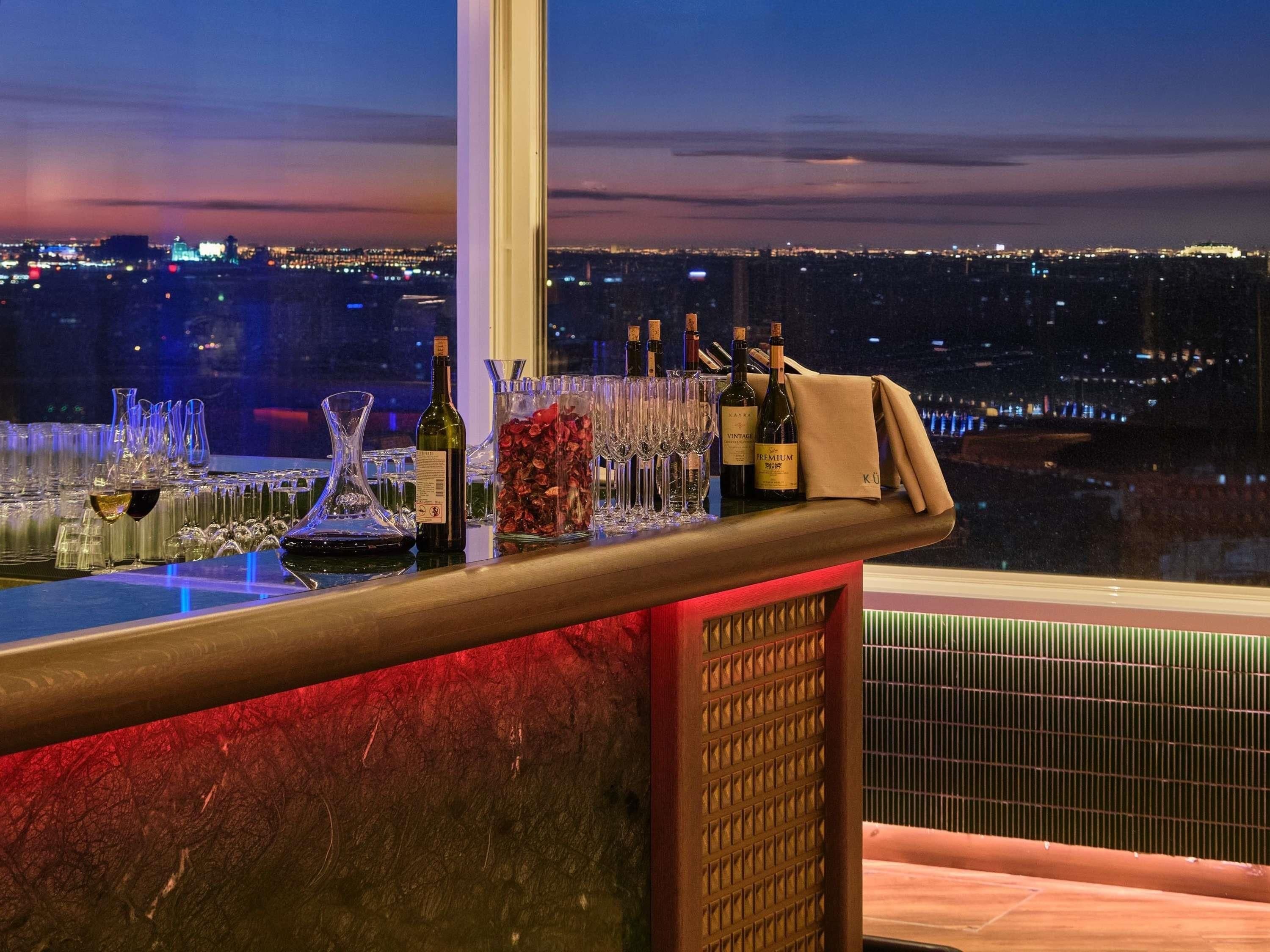 Bar com garrafas e copos, janela com vista para a cidade e balcão iluminado em vermelho.