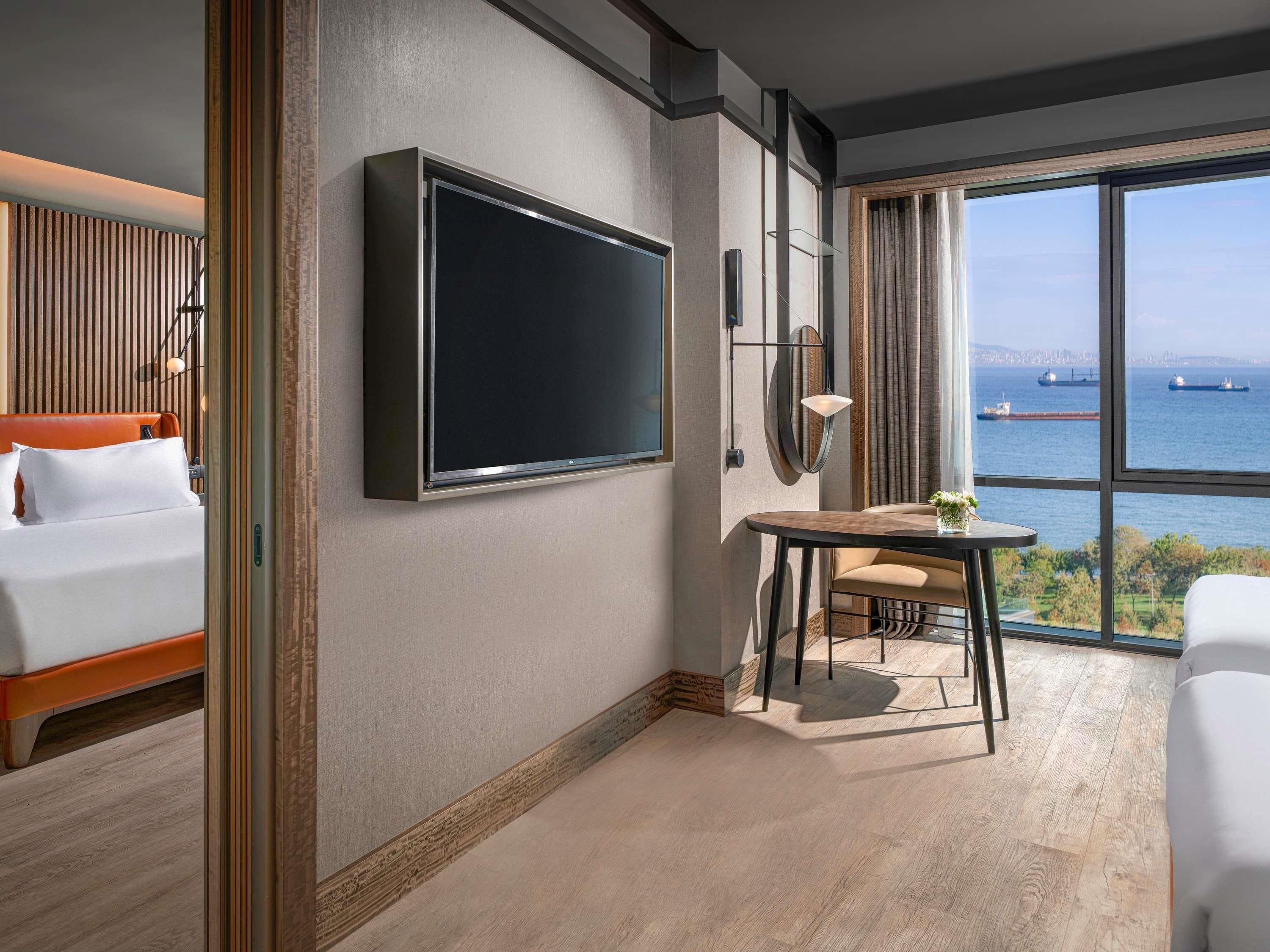 Habitación de hotel con cama, televisor montado en la pared, una mesita con silla y un amplio ventanal con vistas al mar y barcos.