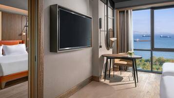 Habitación de hotel con cama, televisor montado en la pared, una mesita con silla y un amplio ventanal con vistas al mar y barcos.