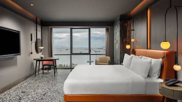 A hotel room with a large bed, a desk, a chair, a TV, and a view of the sea.