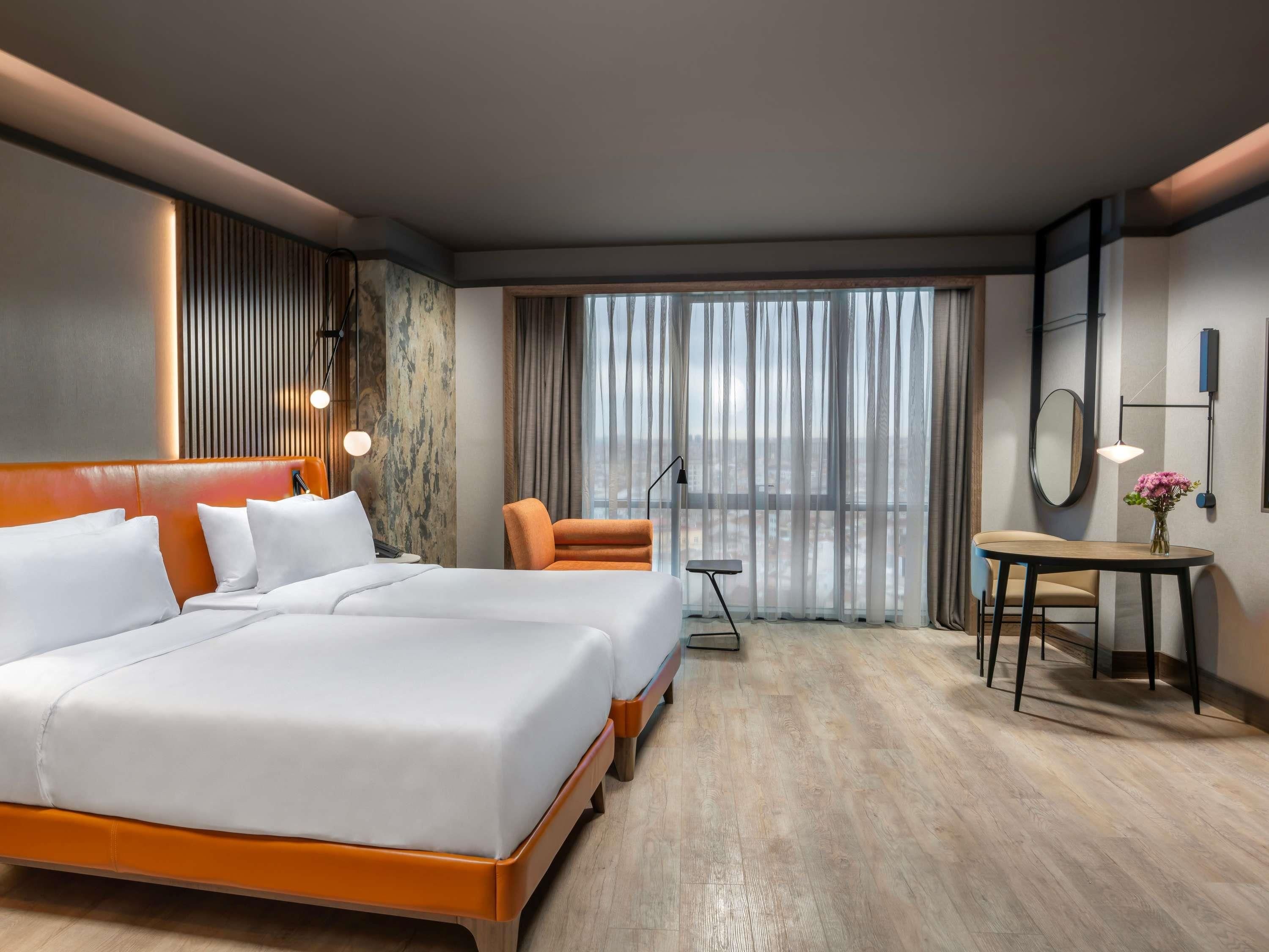Une chambre d’hôtel avec un grand lit, un bureau, une chaise et une vue sur la ville par la fenêtre.
