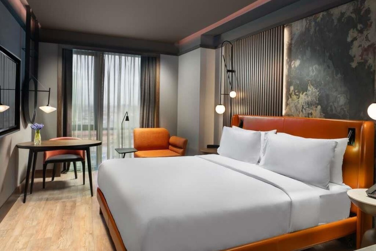 Habitación de hotel con una cama grande, un sillón naranja, una mesita y vistas de la ciudad a través de amplios ventanales.