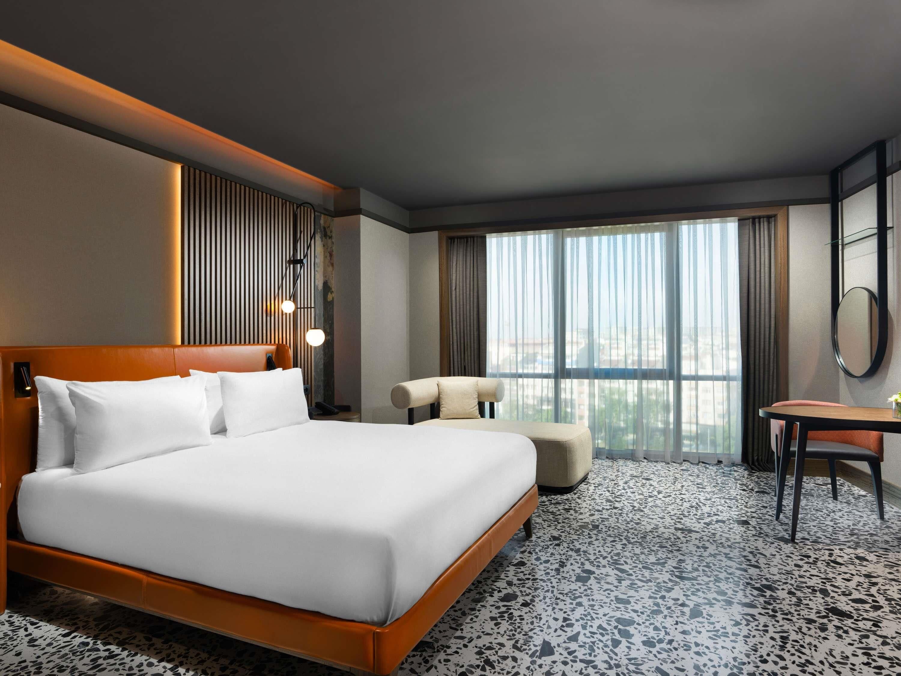A hotel room with a large bed, a desk, a chair, a mirror, and a view of the city through large windows.