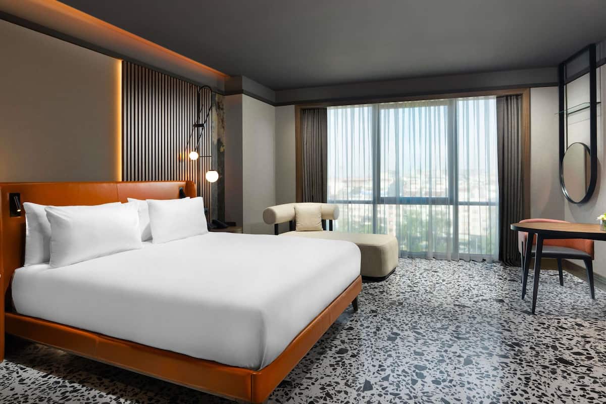 A hotel room with a large bed, a desk, a chair, a mirror, and a view of the city through large windows.