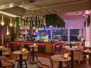 A bar area with a circular ceiling light fixture, a bar counter with various bottles, and seating with round tables and wicker chairs.