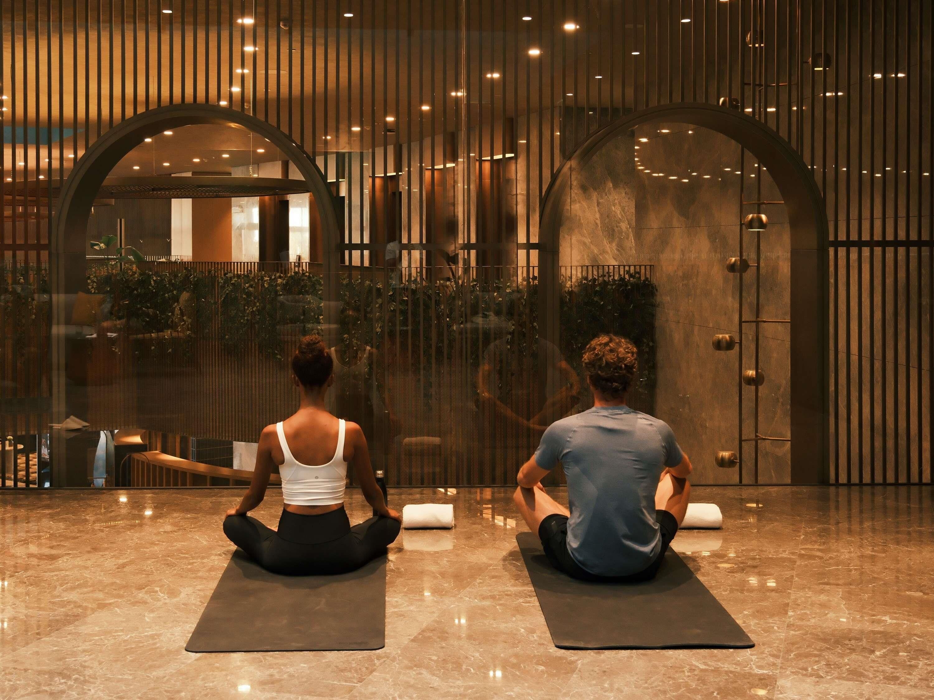 Two individuals seated on yoga mats in a spacious lobby with a modern design, featuring large arched windows and a reflective floor.