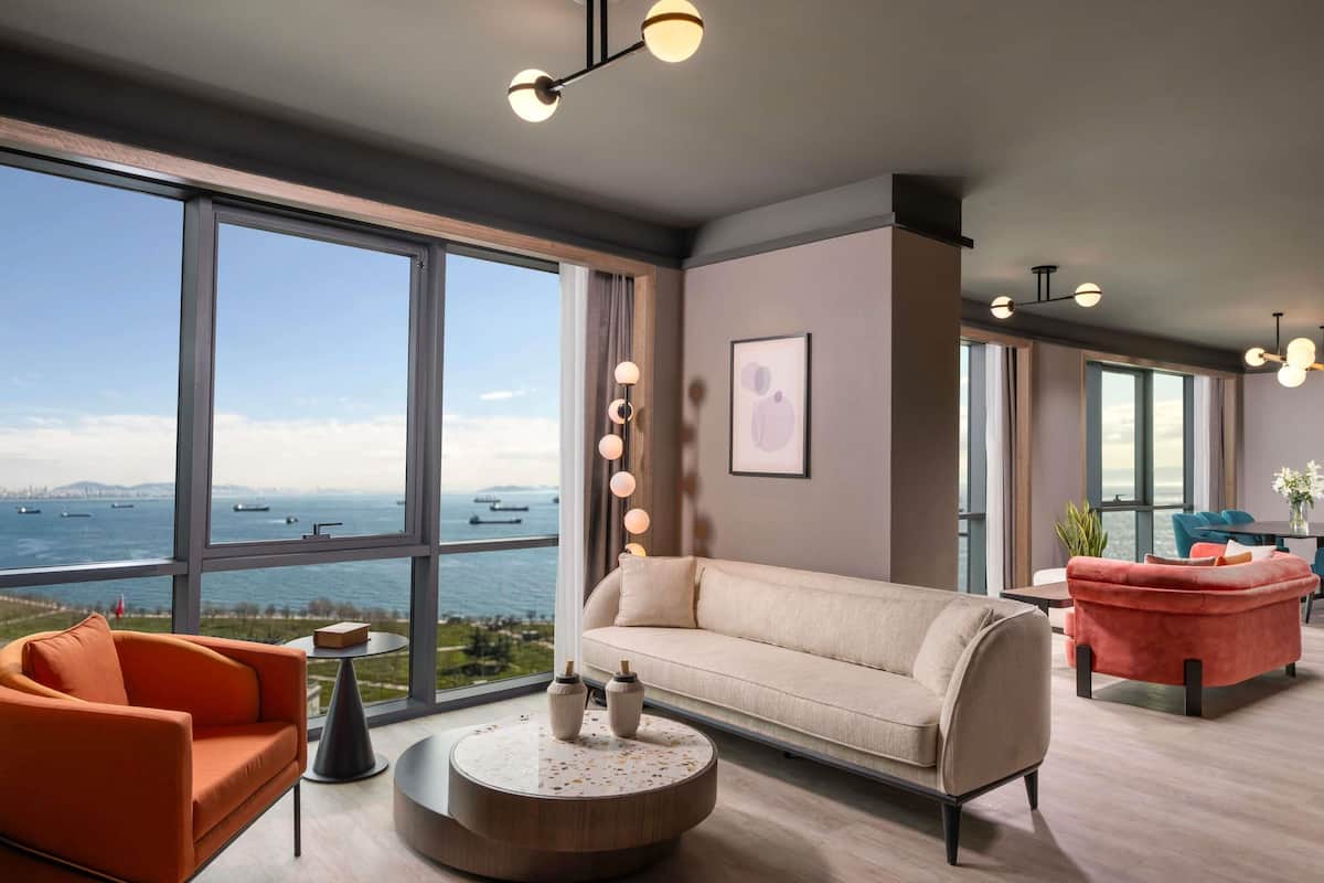 A modern living room with large windows, a beige sofa, an orange armchair, and a red sofa.