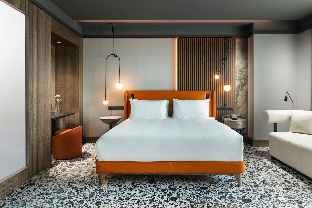 A modern hotel room with a large bed, a small side table, a nightstand, a sofa, and a wall-mounted lamp.
