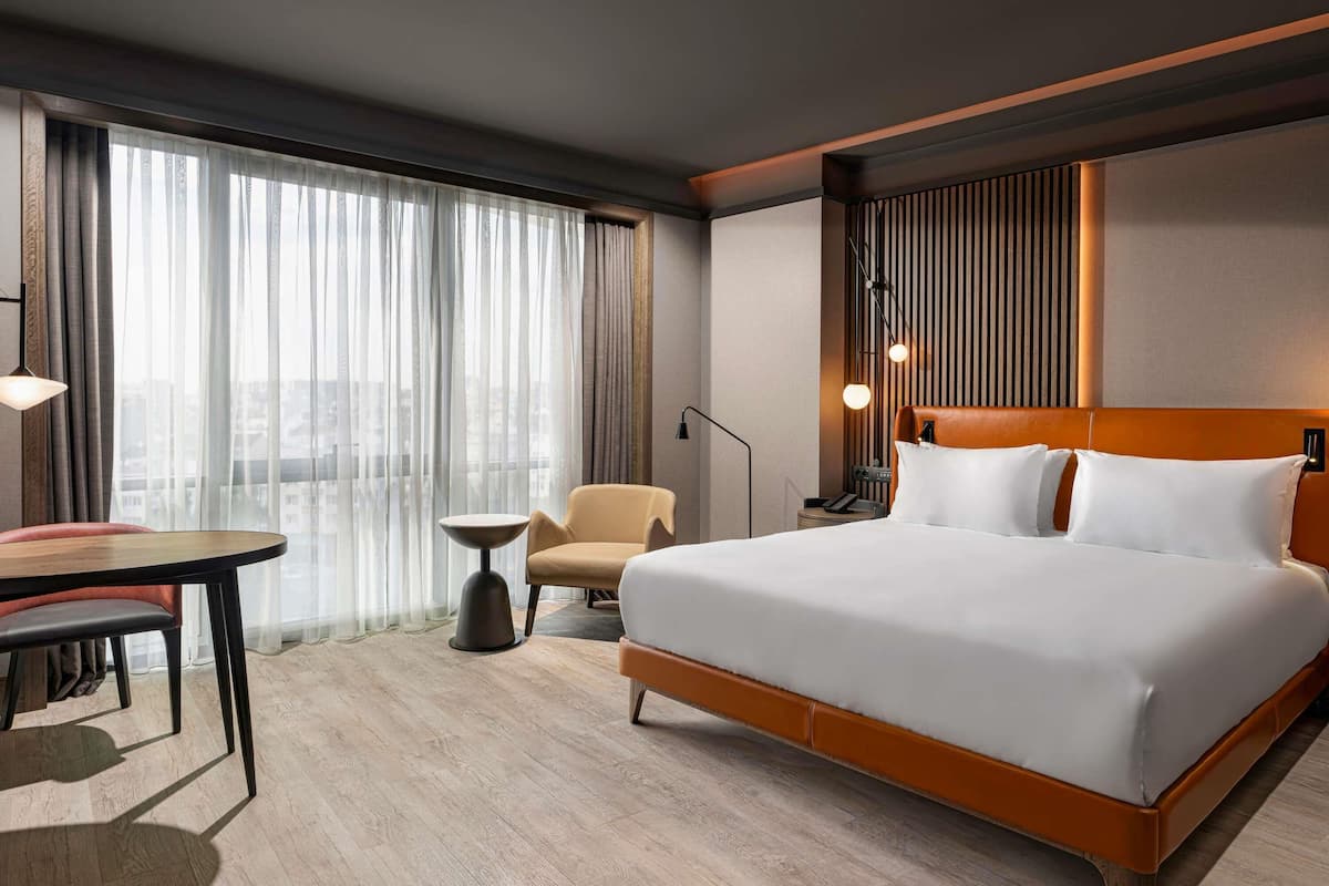A hotel room with a large bed, a desk, a chair, and a view of the city through sheer curtains.