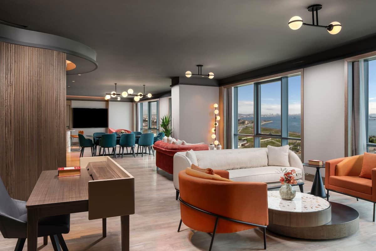 A modern living room with a wooden accent wall, a dining area with teal chairs, and a large window offering a city view.