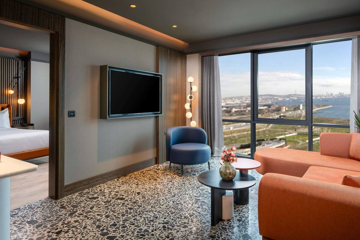 A hotel room with a bed, a TV mounted on the wall, a seating area with a chair and sofa, a small table, and a view of the city through large windows.