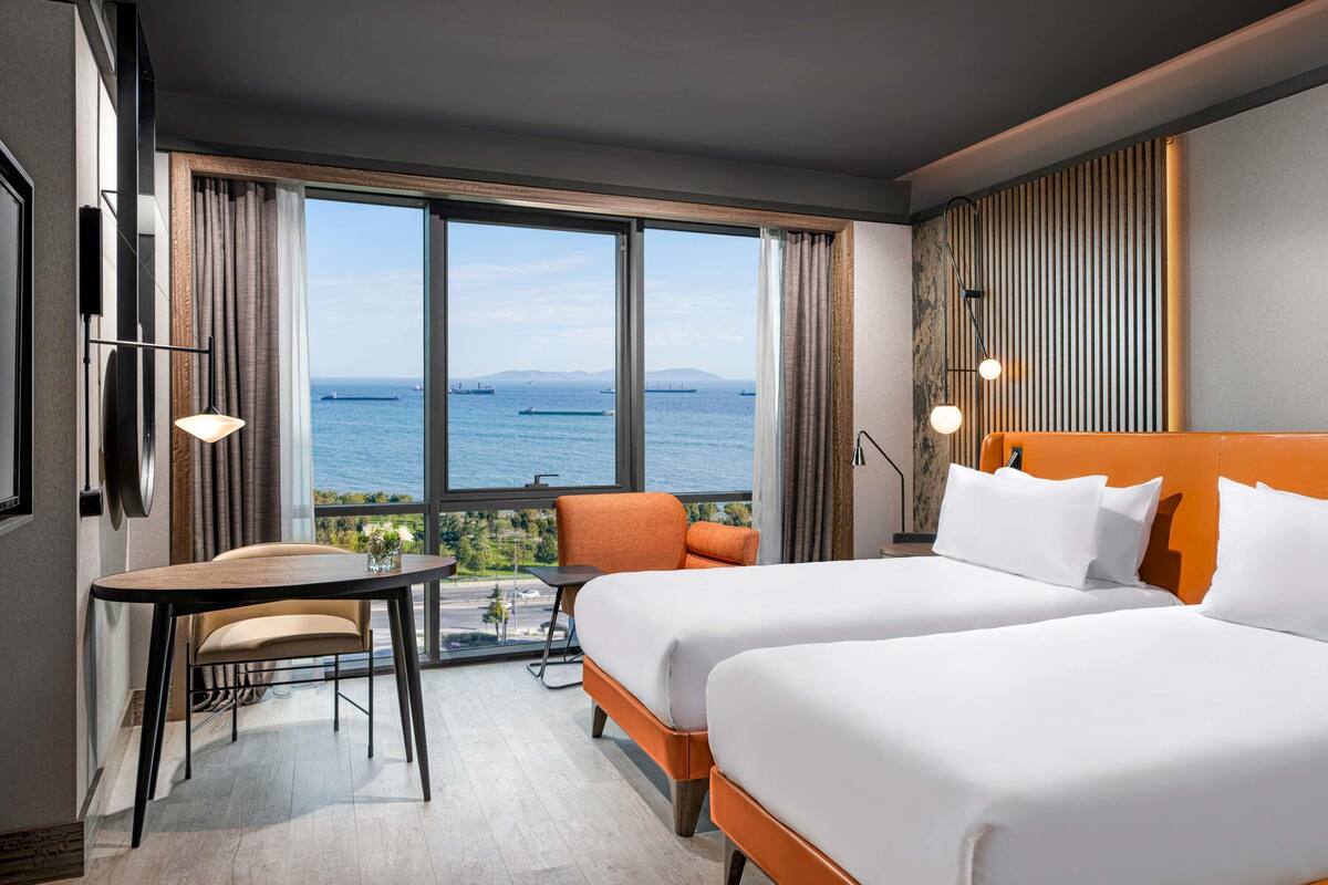 A hotel room with two beds, a small table, a chair, and a view of the sea.