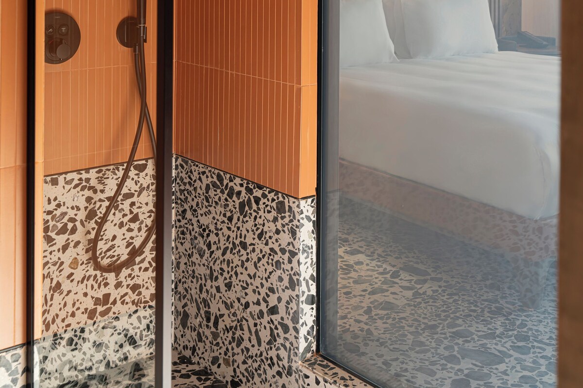 Un baño moderno con ducha, separador de vidrio y cama con ropa de cama blanca.