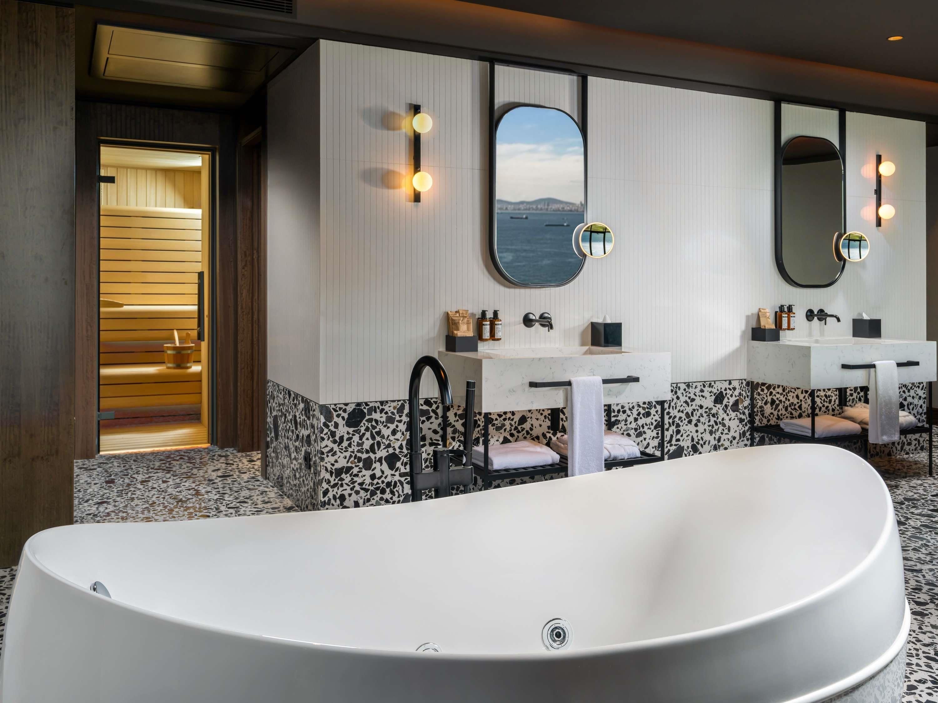 Un baño moderno con una bañera independiente, dos lavamanos con espejos y una sauna visible a través de una puerta abierta.