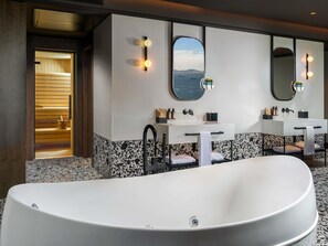 Ein modernes Badezimmer mit freistehender Badewanne, zwei Waschbecken mit Spiegeln und einer Sauna, die durch eine geöffnete Tür sichtbar ist.
