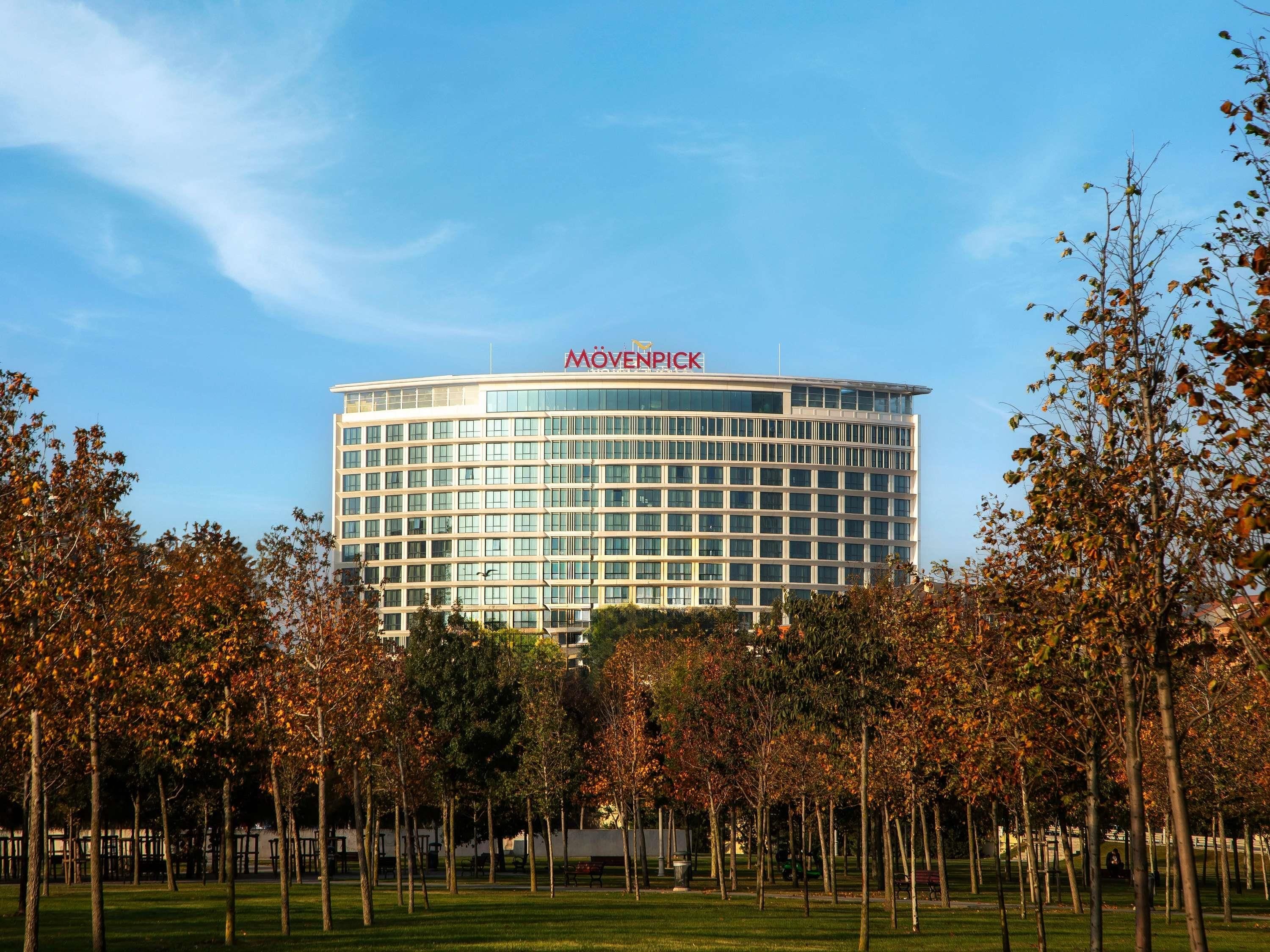 Un moderno edificio hotel con cielo sereno, circondato da un parco con alberi e panchine.