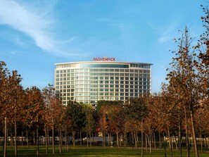 Un hotel moderno con cielo despejado, rodeado por un parque con árboles y bancos.