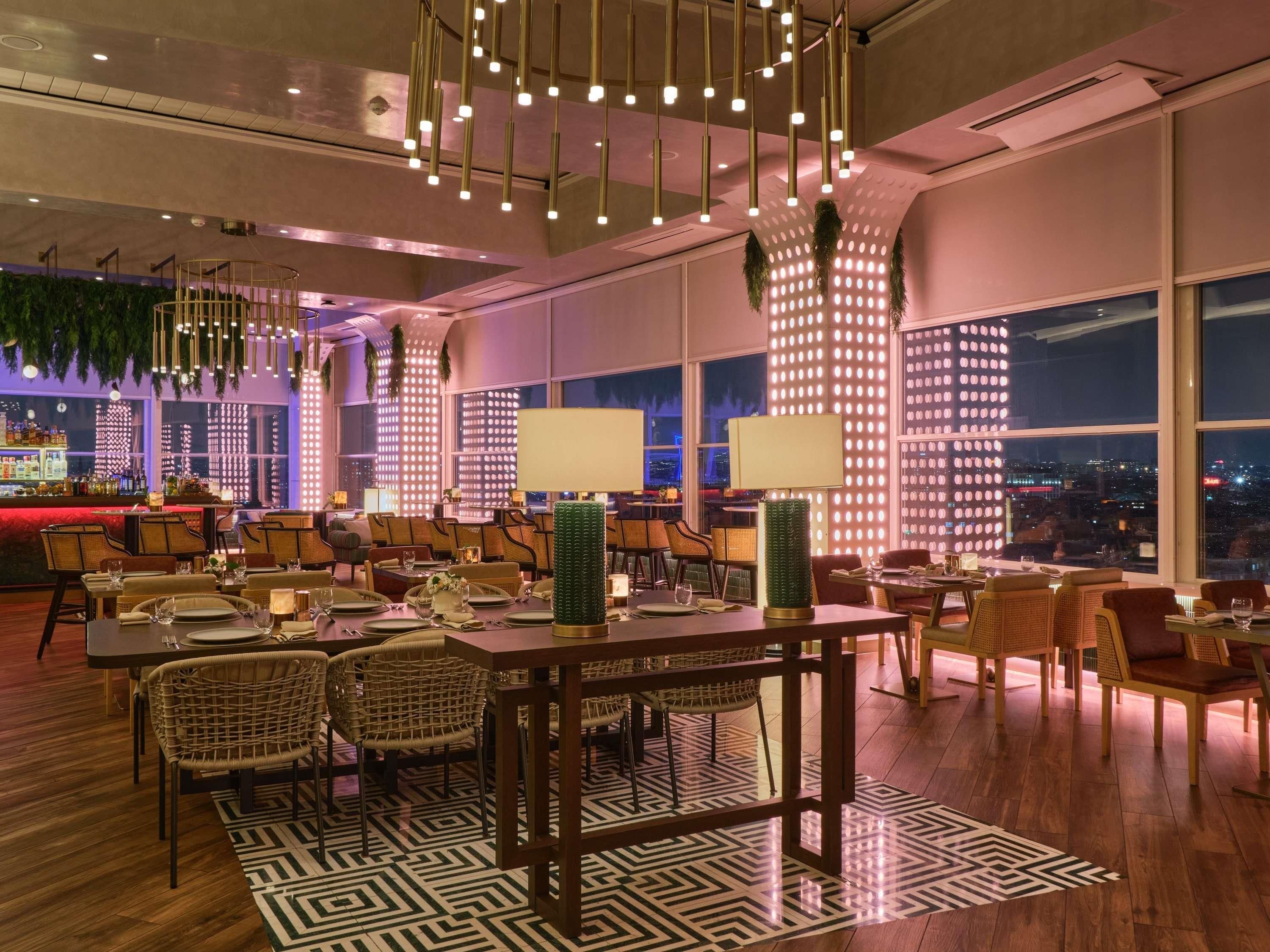 A modern restaurant with high ceilings, hanging lights, and a bar area with a city view.