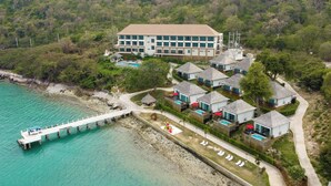 An aerial view of a resort with a large building, multiple guest houses, a swimming pool, and a pier extending into turquoise waters. - Novotel Koh Si Chang (Ko Sichang)
