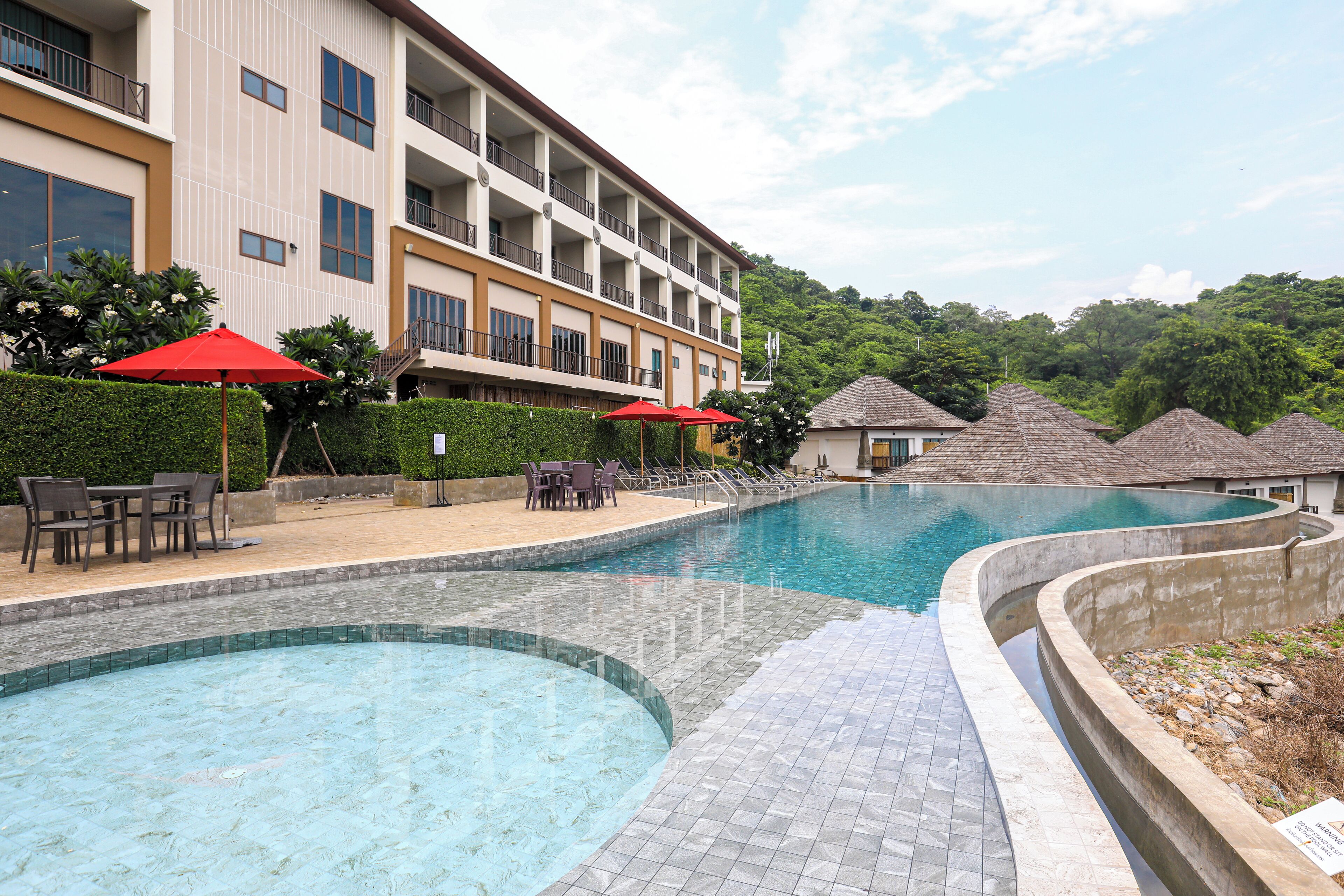Ein Schwimmbecken mit bogenförmigem Design, umgeben von einer gefliesten Terrasse. Rote Sonnenschirme und Sitzmöbel sind vorhanden.