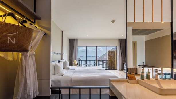 A hotel room with a large bed, a balcony with a view, a bathroom with a sink and toiletries, and a hanging basket. - Novotel Koh Si Chang (Ko Sichang)