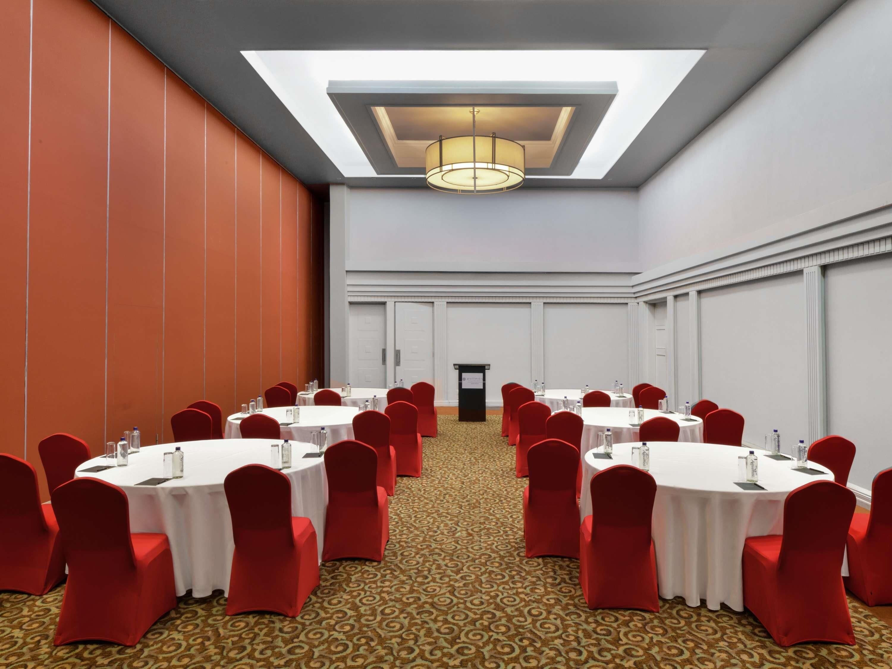 Un salón de conferencias con mesas redondas, sillas rojas y una alfombra estampada.
