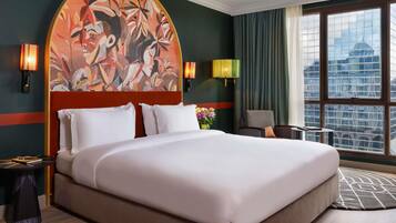 Habitación de hotel con una cama grande, cabecera con un mural floral, dos apliques de pared, vista de la ciudad a través de amplios ventanales y una mesita de noche con lámpara.
