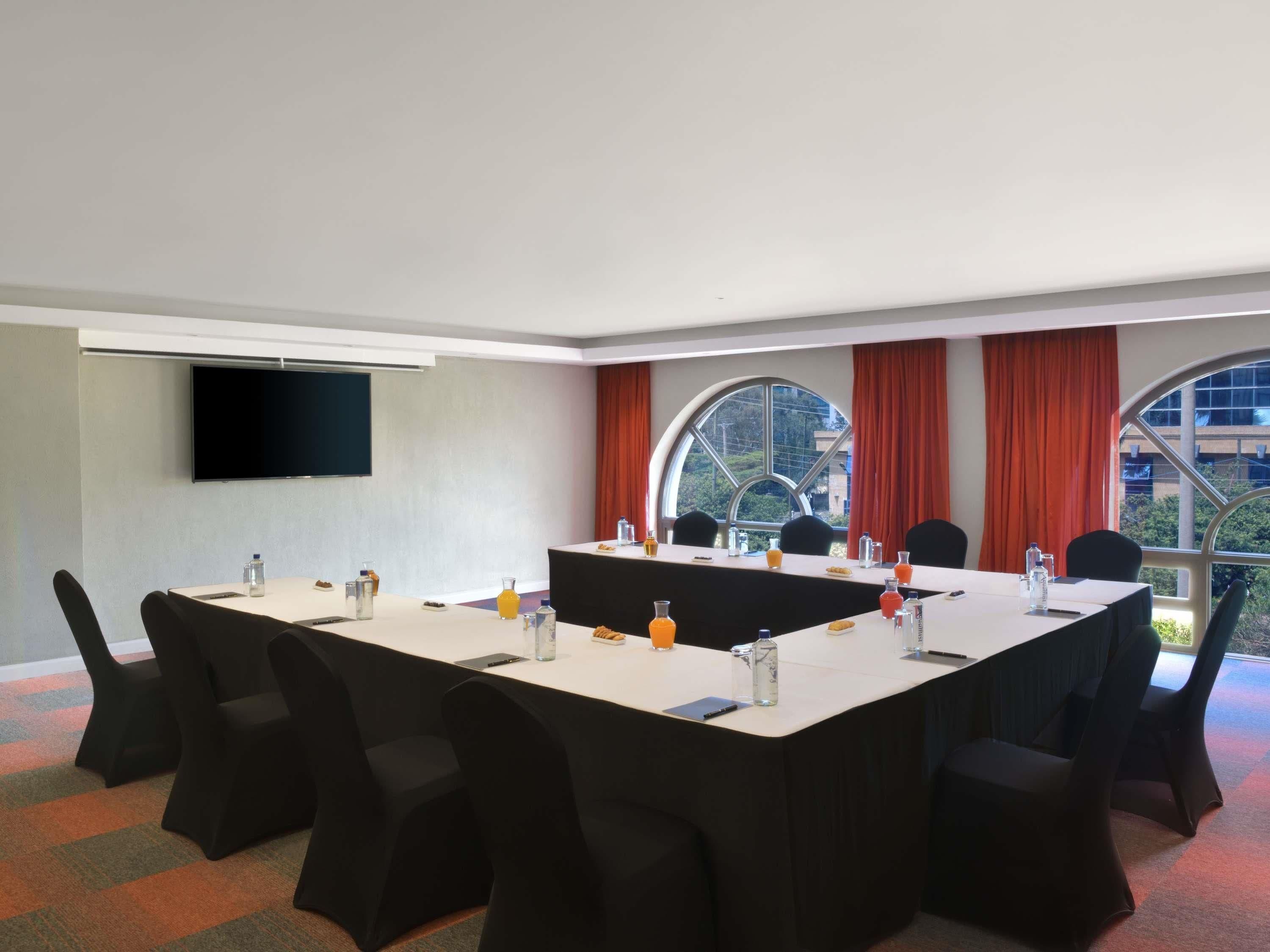 A conference room with a long table, chairs, a flat-screen TV, and large windows with curtains.