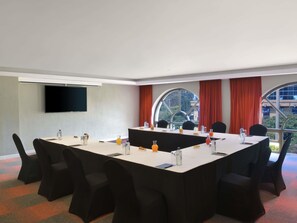 Un salón de conferencias con una larga mesa, sillas, un televisor de pantalla plana y amplias ventanas con cortinas.