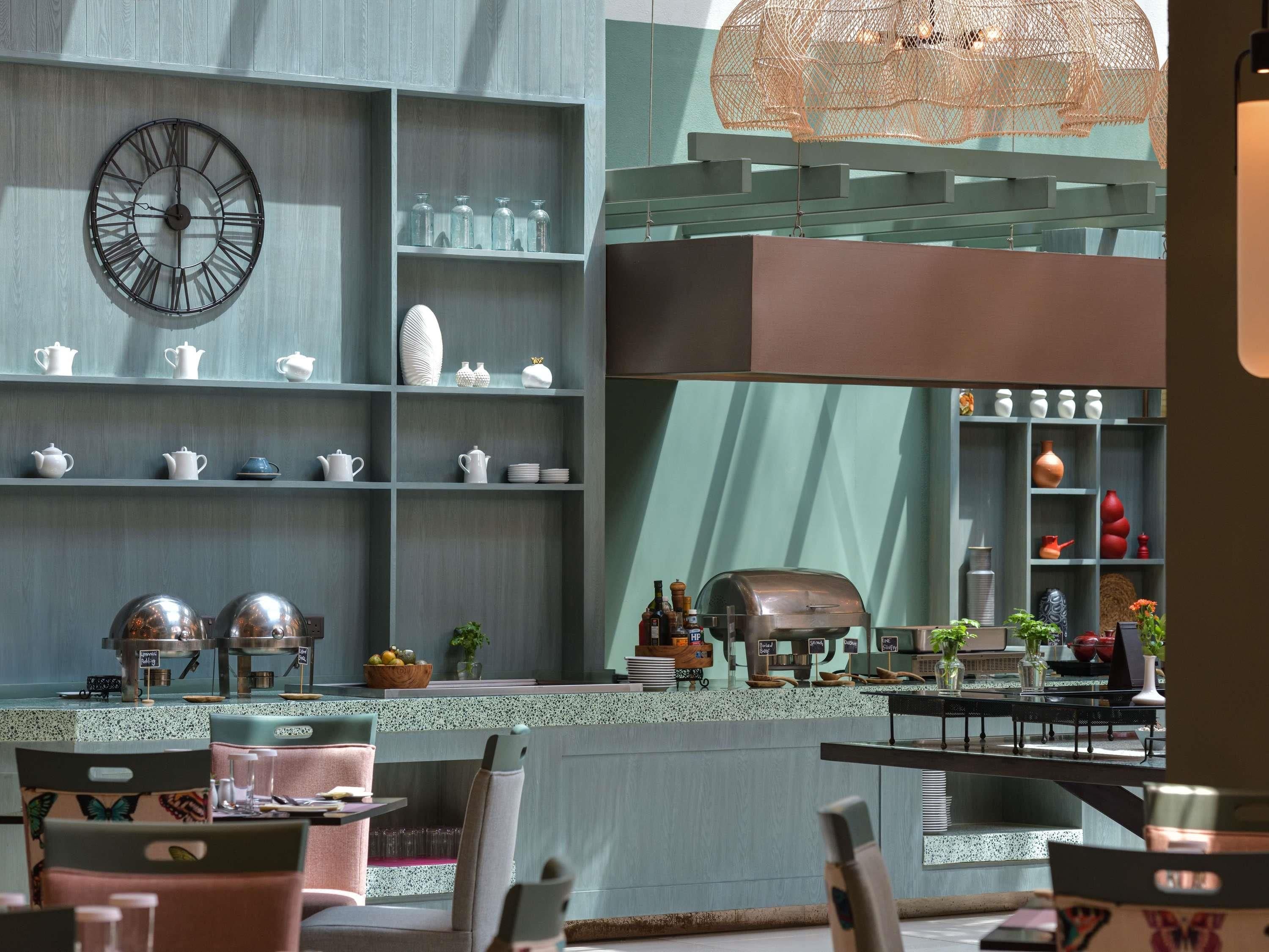 A modern kitchen with a large clock, shelves with kitchenware, a grill, and a dining area with chairs and tables.