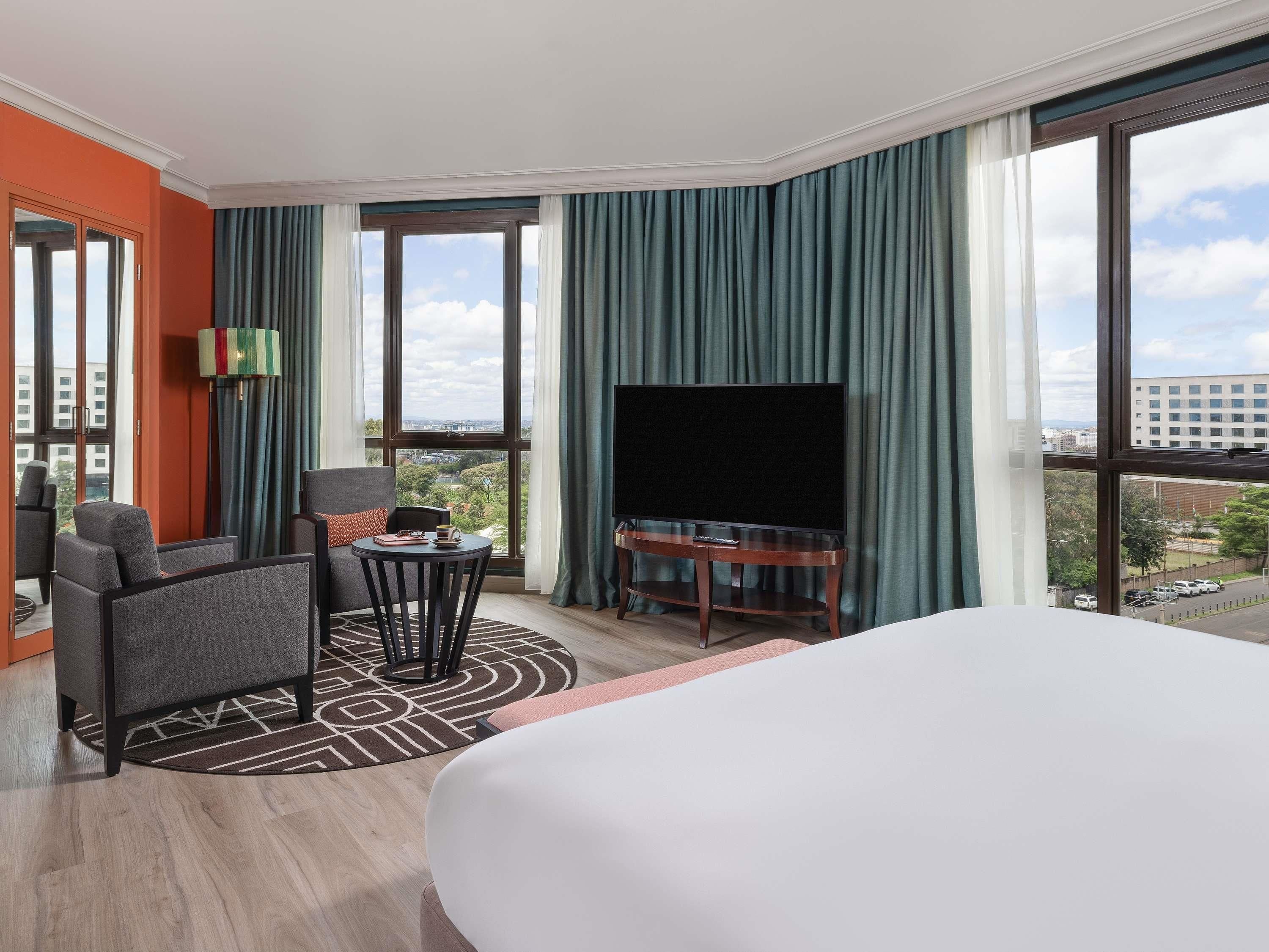 A hotel room with a large bed, a TV on a wooden stand, a seating area with a table and chairs, and large windows offering a city view.