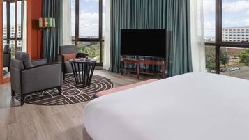 A hotel room with a large bed, a TV on a wooden stand, a seating area with a table and chairs, and large windows offering a city view.