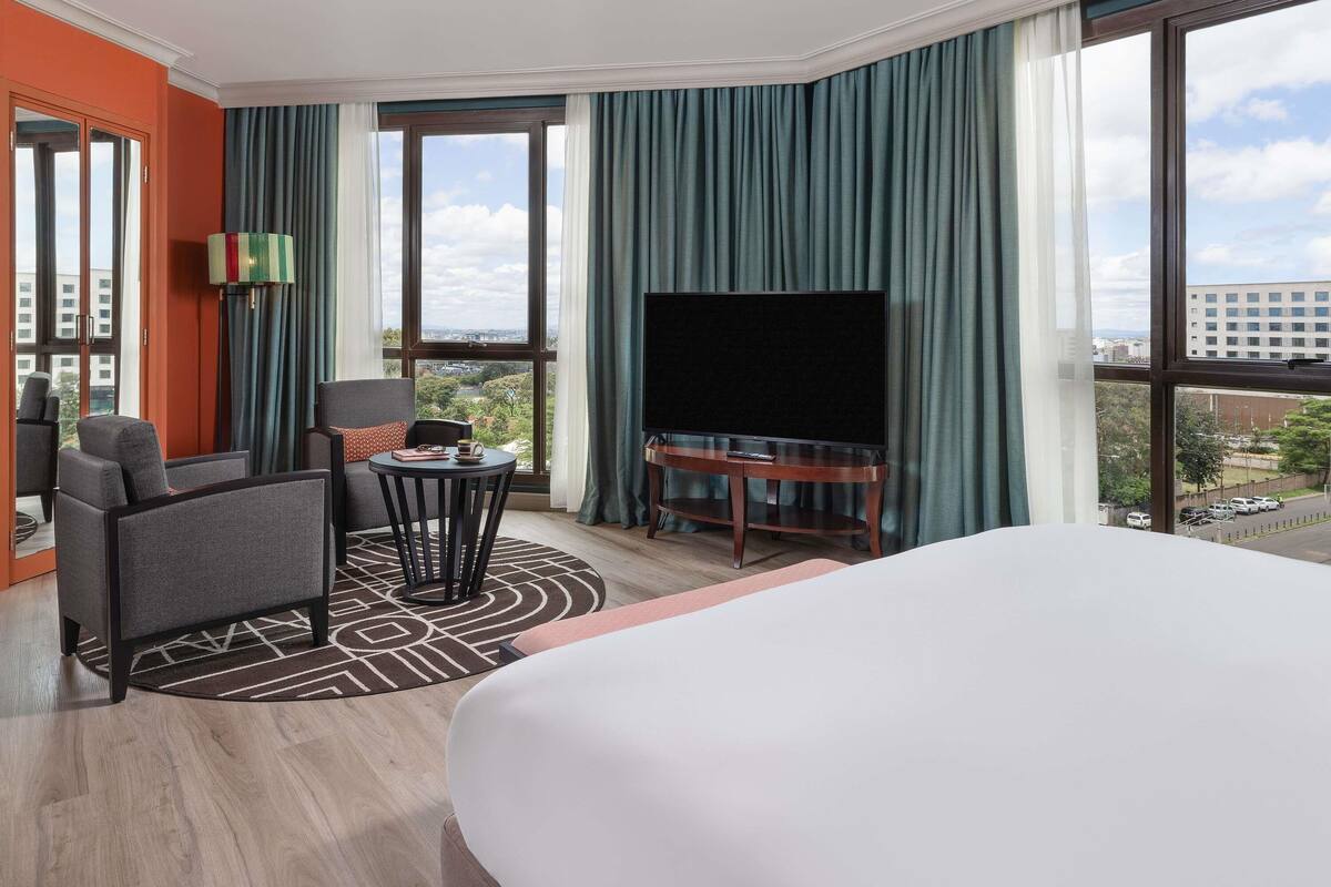 A hotel room with a large bed, a TV on a wooden stand, a seating area with a table and chairs, and large windows offering a city view.