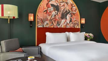 A hotel room with a large bed, a chair, a small table with a cup and notebook, and a wall painting.