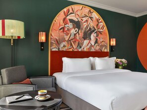 Habitación de hotel con una cama grande, una silla, una mesita con una taza y un cuaderno, y un cuadro en la pared.