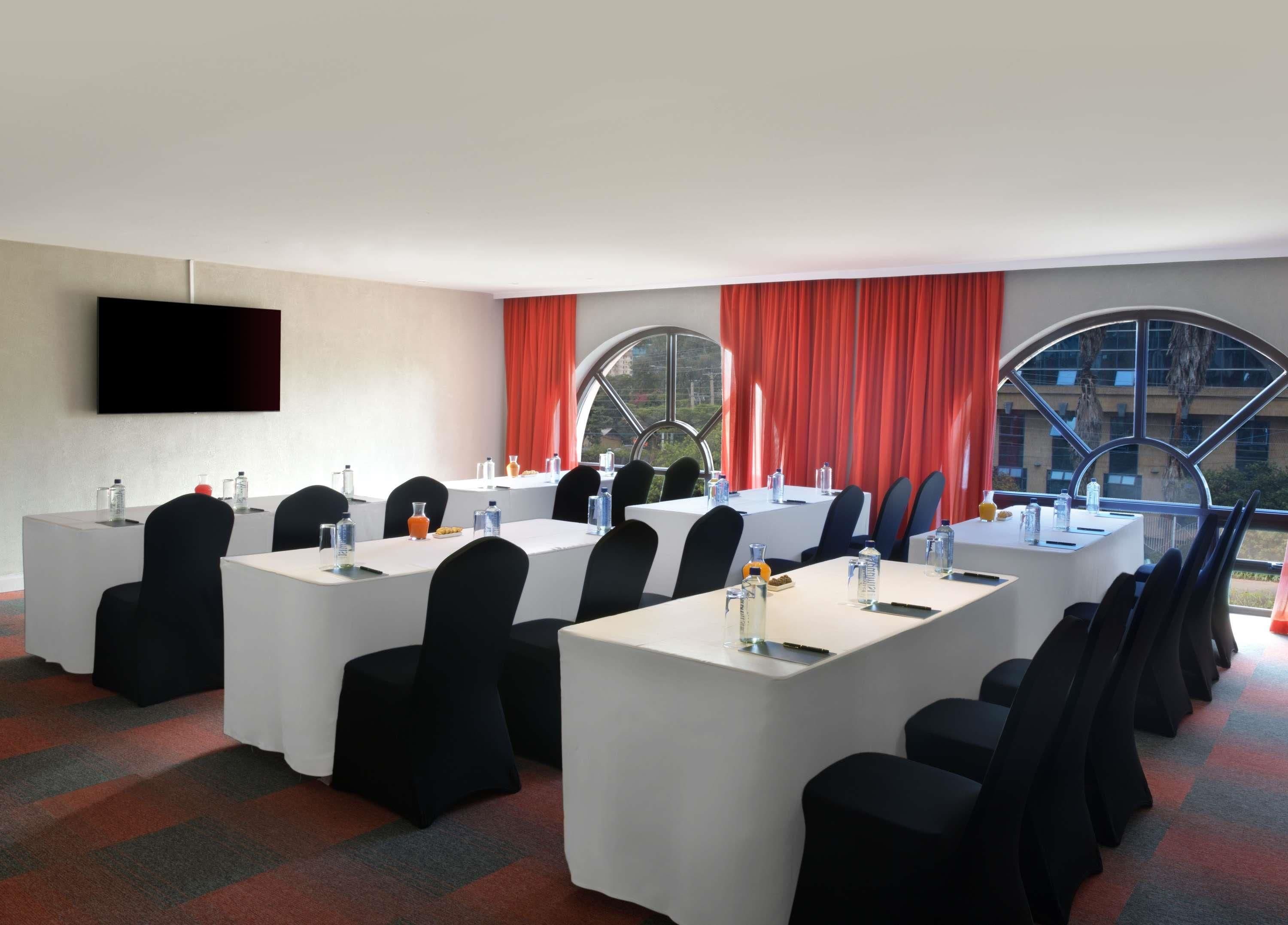 Un salón de conferencias con varias mesas, sillas y un amplio ventanal con cortinas rojas.