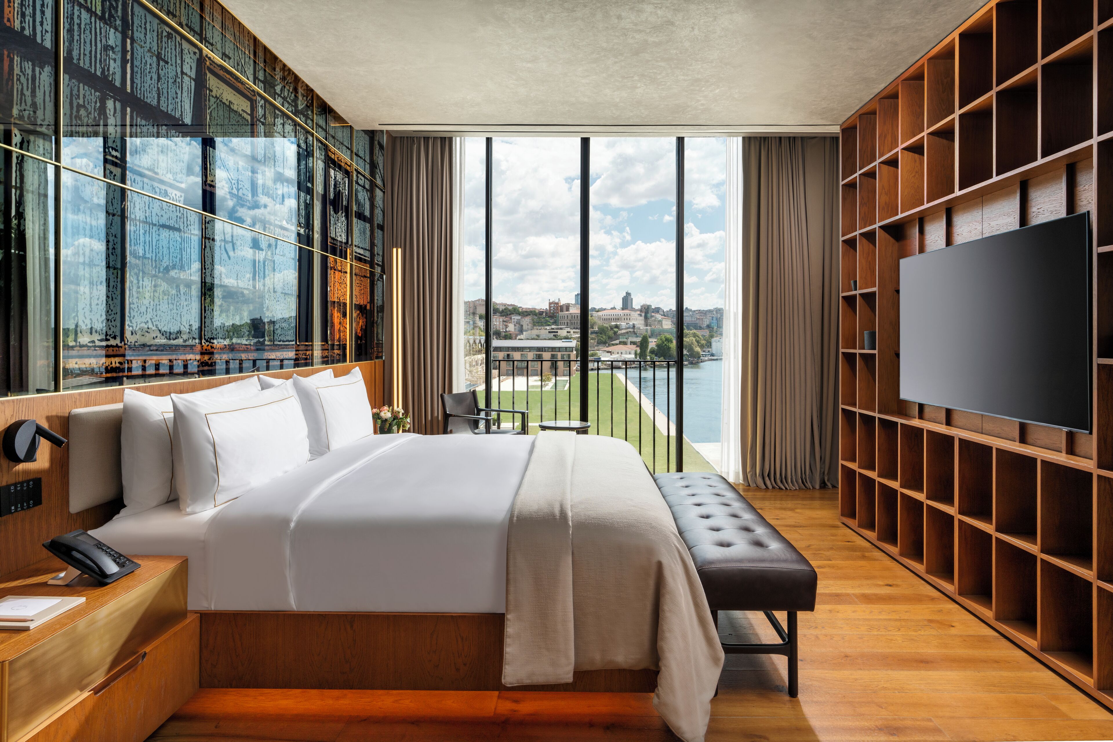A modern hotel room with a large bed, a TV, a balcony with a city view, and a wooden bookshelf.