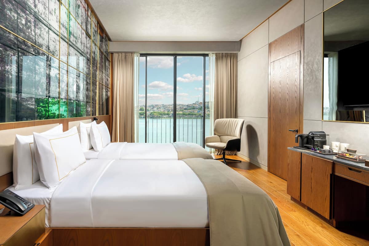 Habitación de hotel con una cama grande, un escritorio con cafetera, una silla y vistas a una masa de agua a través de amplias ventanas.