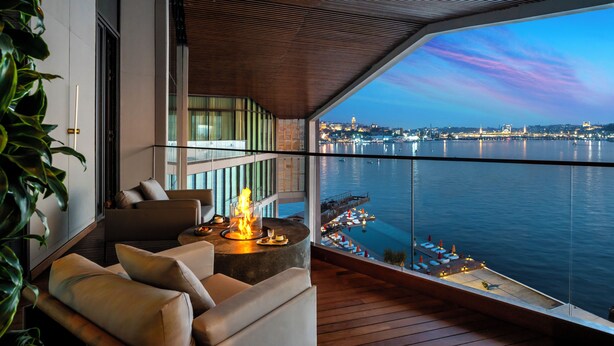 Un balcon moderne offrant une vue sur l’eau et le paysage urbain, avec un canapé, une table ronde dotée d’un foyer et une rambarde en verre.