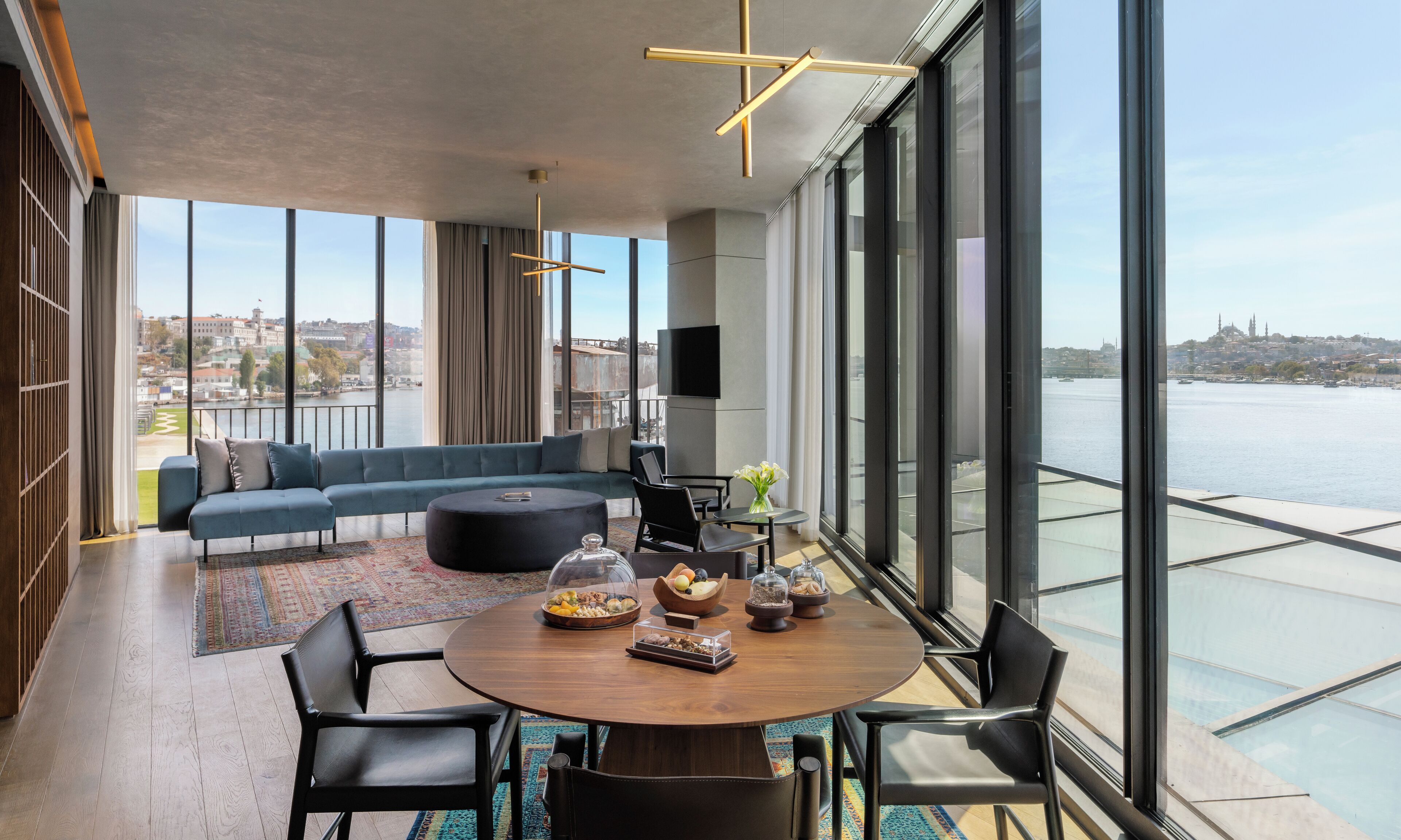 A modern living room with a large sofa, a round dining table, and a view of a river and cityscape.