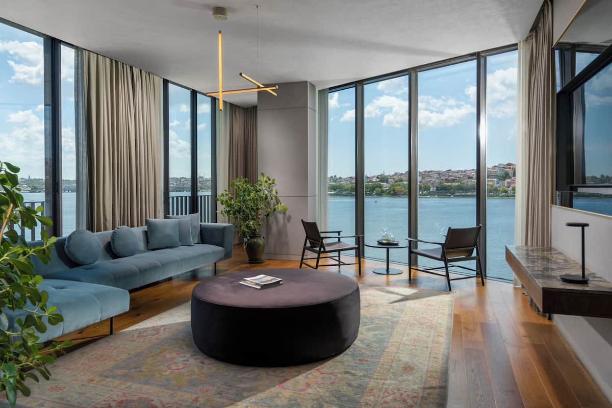Una sala de estar moderna con un sofá grande, un otoman redonda y una mesita. Hay dos sillones y una planta en maceta. La habitación tiene ventanas amplias con vistas a un cuerpo de agua y edificios.