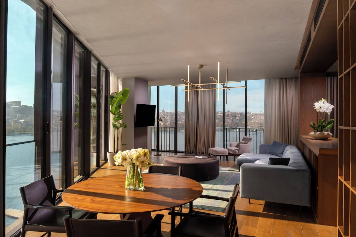 Una sala de estar moderna con una gran mesa de madera, asientos cómodos y vistas de un paisaje urbano a través de ventanas del piso al techo.