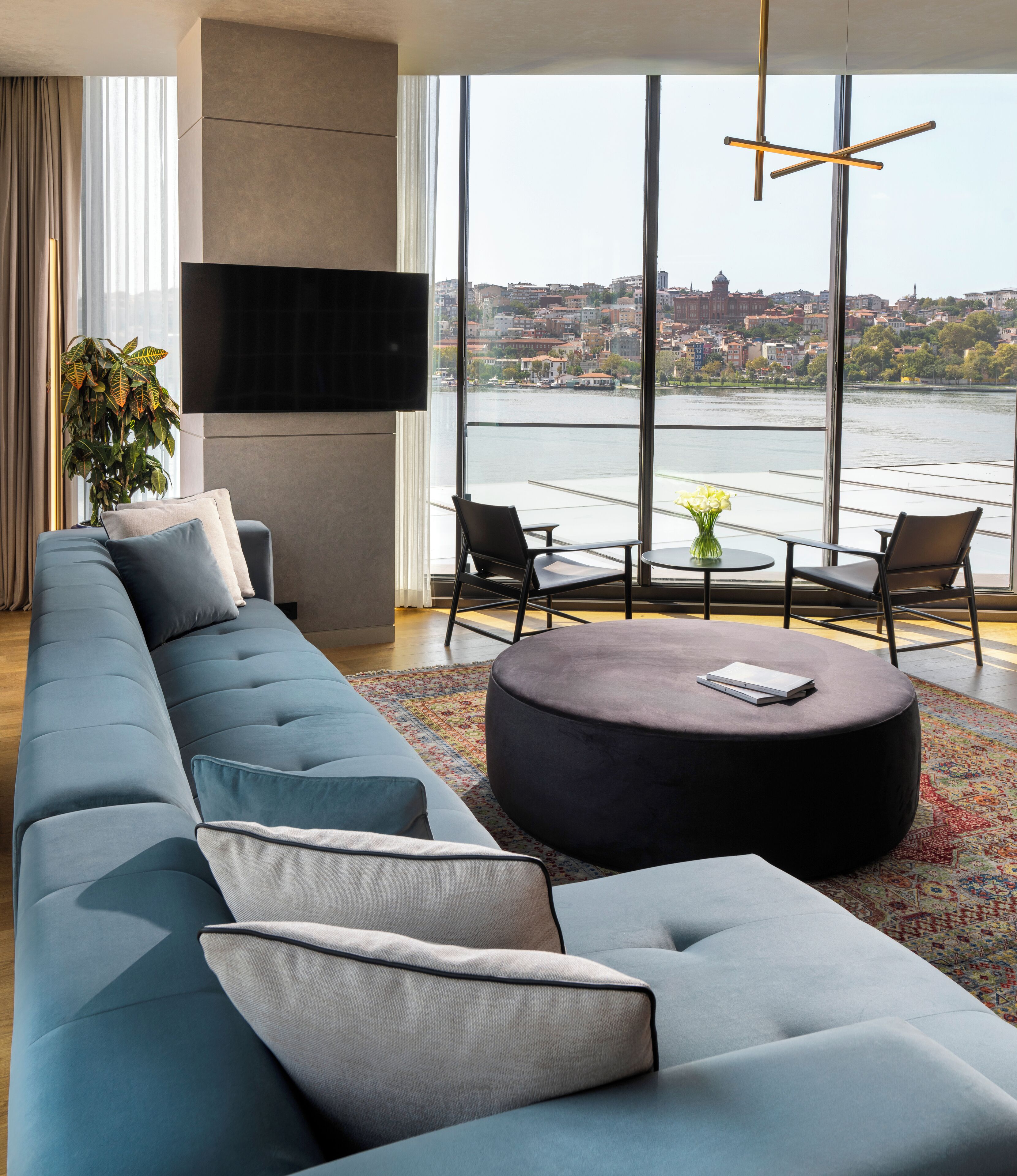 A modern living room with a large sectional sofa, a round ottoman, and a view of a cityscape through large windows.